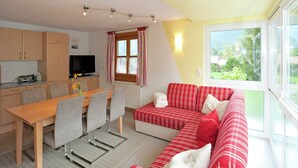 Un salon chaleureux avec un canapé à carreaux rouges et blancs, une table à manger avec des chaises, et un coin cuisine doté de meubles en bois et d’un téléviseur.