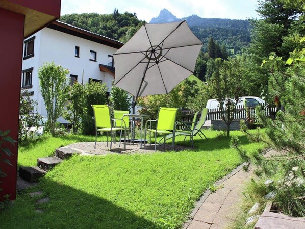 A patio with a table and chairs, a large umbrella, and a view of a white building and greenery.