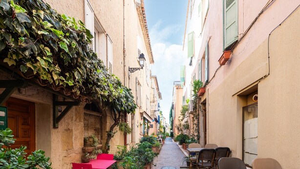 Un callejón estrecho con un camino de ladrillo, sillas y mesas rojas a un lado, y un edificio con contraventanas verdes y plantas trepadoras al otro.