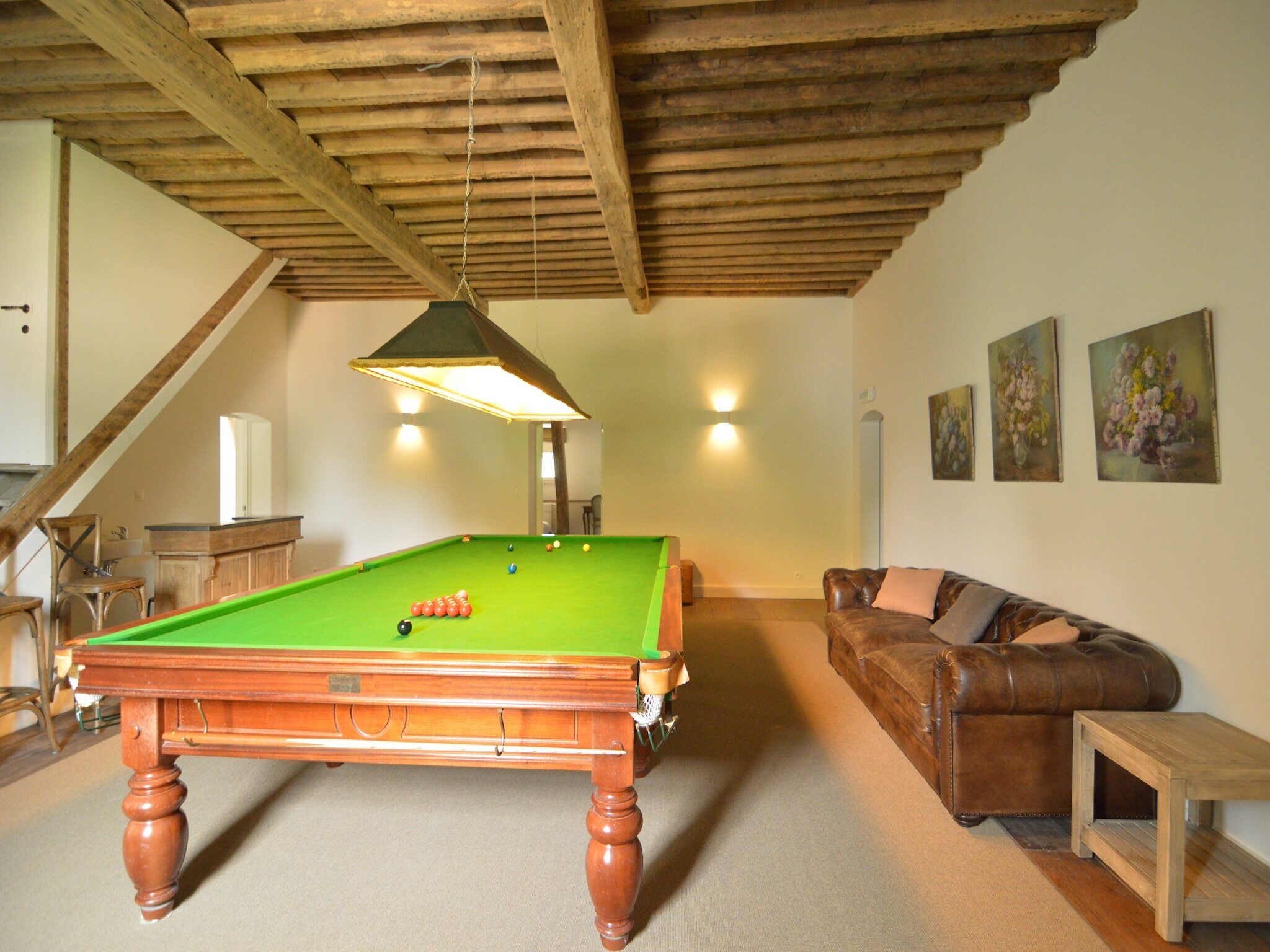 Une table de billard dans une pièce dotée d’un plafond en bois, d’un canapé en cuir marron et d’une petite table en bois.