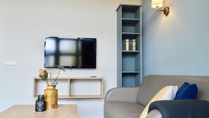 Una sala de estar con un televisor de pantalla plana montado en la pared, una mesa de centro de madera con jarrones decorativos, un sofá gris con cojines y un estante azul adosado a la pared.