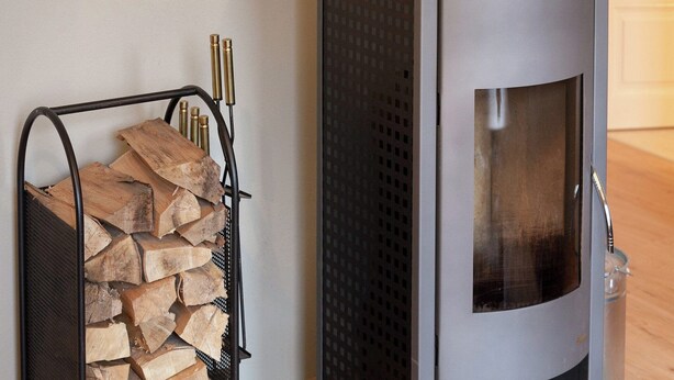 Un poêle à bois moderne dot é d’une porte en verre, placé à côté d’un support métallique où est entreposé le bois de chauffage.