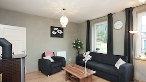 Un salon moderne avec deux canapés sombres, une table basse en bois, un lustre, une horloge et une vue sur l’extérieur grâce à de grandes fenêtres.