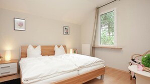 Une chambre à coucher avec un lit en bois, du linge de lit blanc, une table de chevet avec une lampe et une fenêtre donnant sur des arbres.