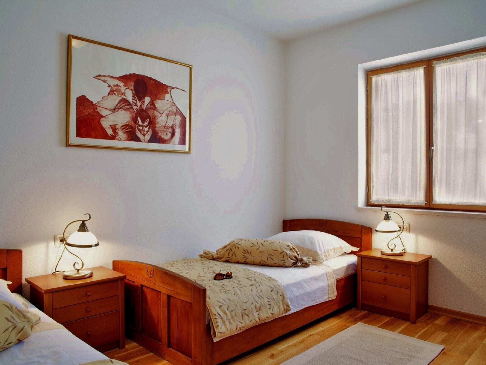 A bedroom with a wooden bed, two bedside tables with lamps, a window with curtains, and a framed picture on the wall.