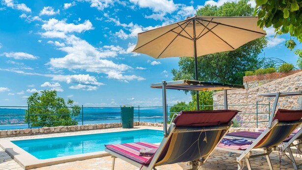 A poolside area with lounge chairs and an umbrella, overlooking a clear blue sky and a distant view of the sea.