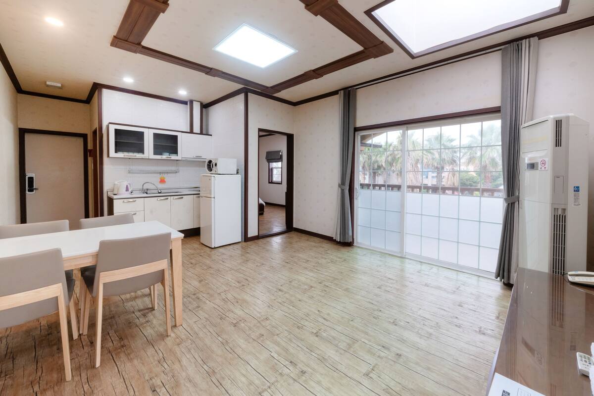 A modern kitchen with white cabinets, a refrigerator, and a microwave. There is a dining area with a table and chairs. The room has wooden flooring and a large window with curtains.