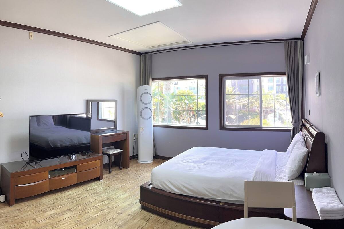 A modern hotel room with a large bed, a TV, a wooden dresser, and a window with a view of greenery.