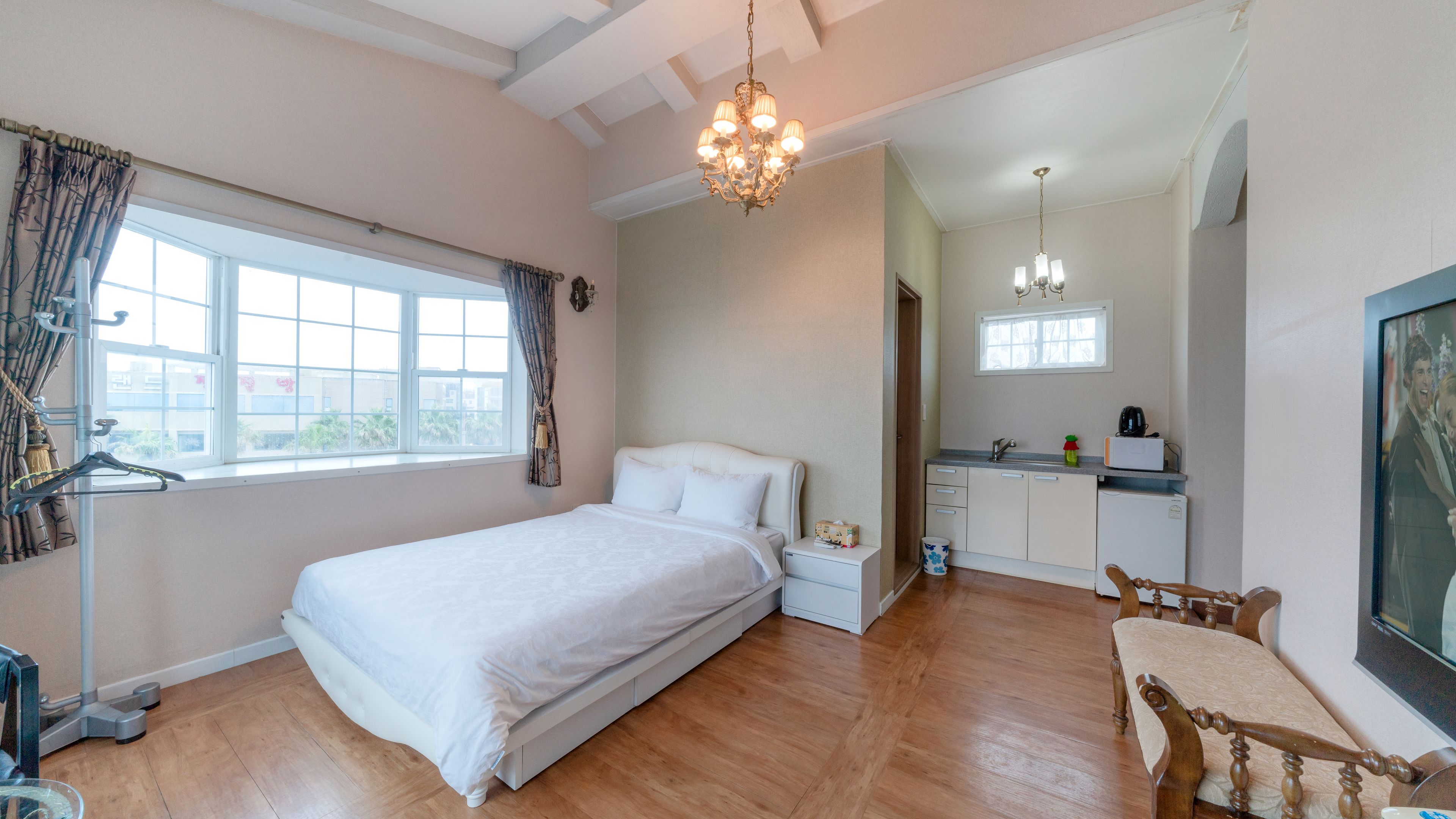 A bedroom with a large bed, a small bedside table, a window with curtains, a wooden floor, and a kitchen area with a sink and cabinets.