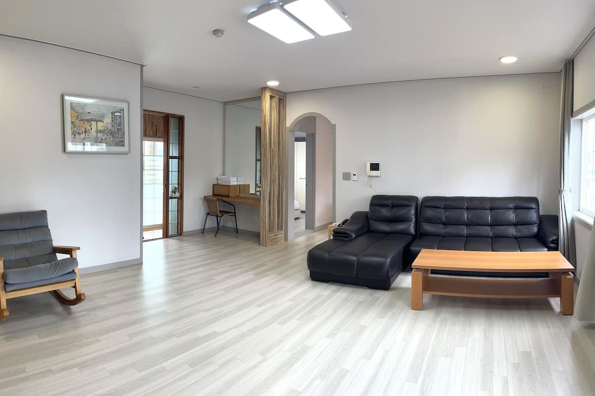 A spacious living area with a wooden floor, a black leather sofa, a wooden coffee table, and a grey rocking chair.