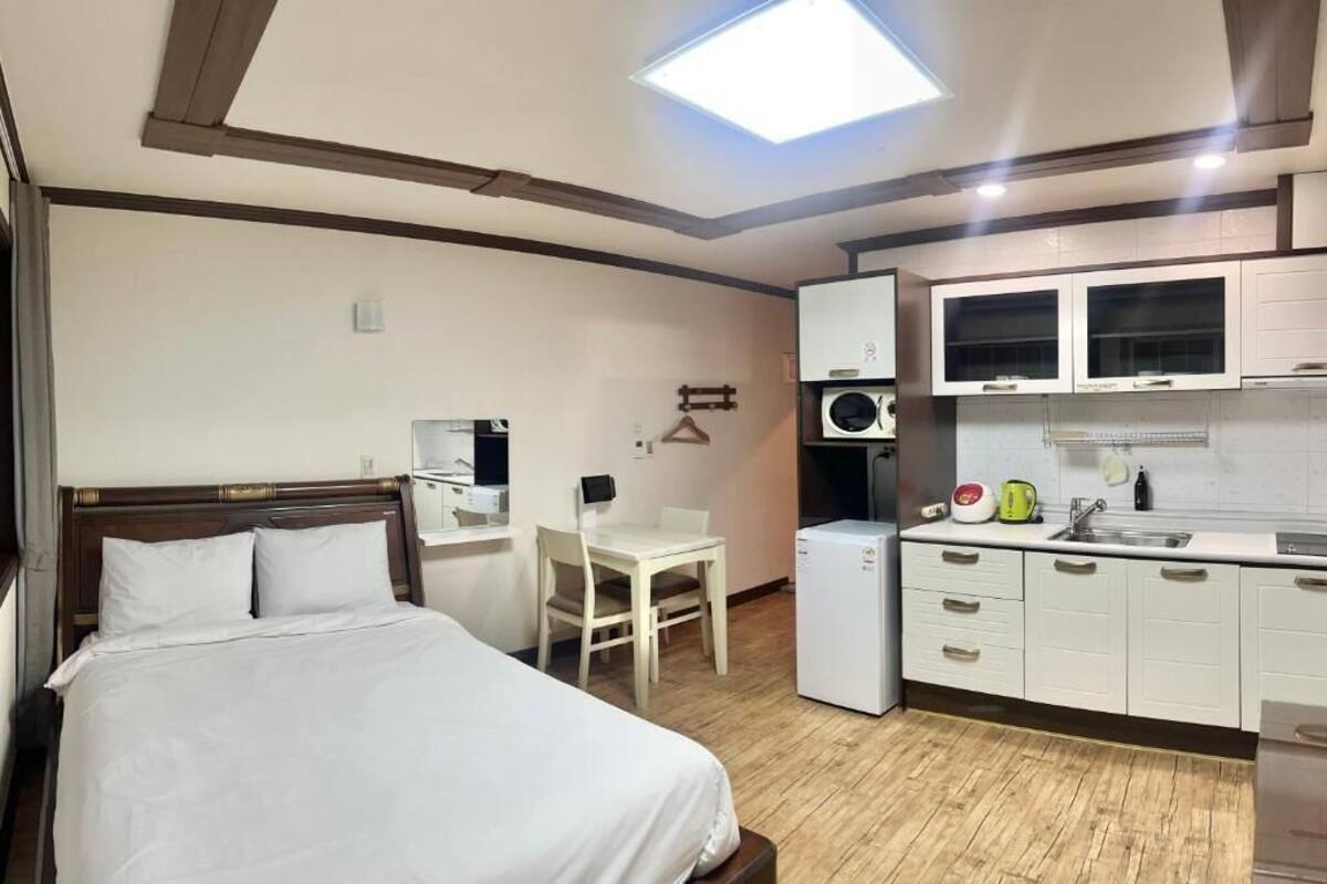 A compact hotel room with a bed, a small desk, a kitchenette, and a ceiling light.