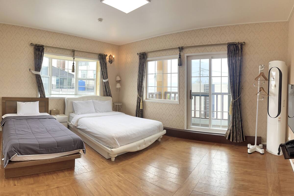 A hotel room with two beds, a large window with curtains, a ceiling light, and a bedside table with a lamp.