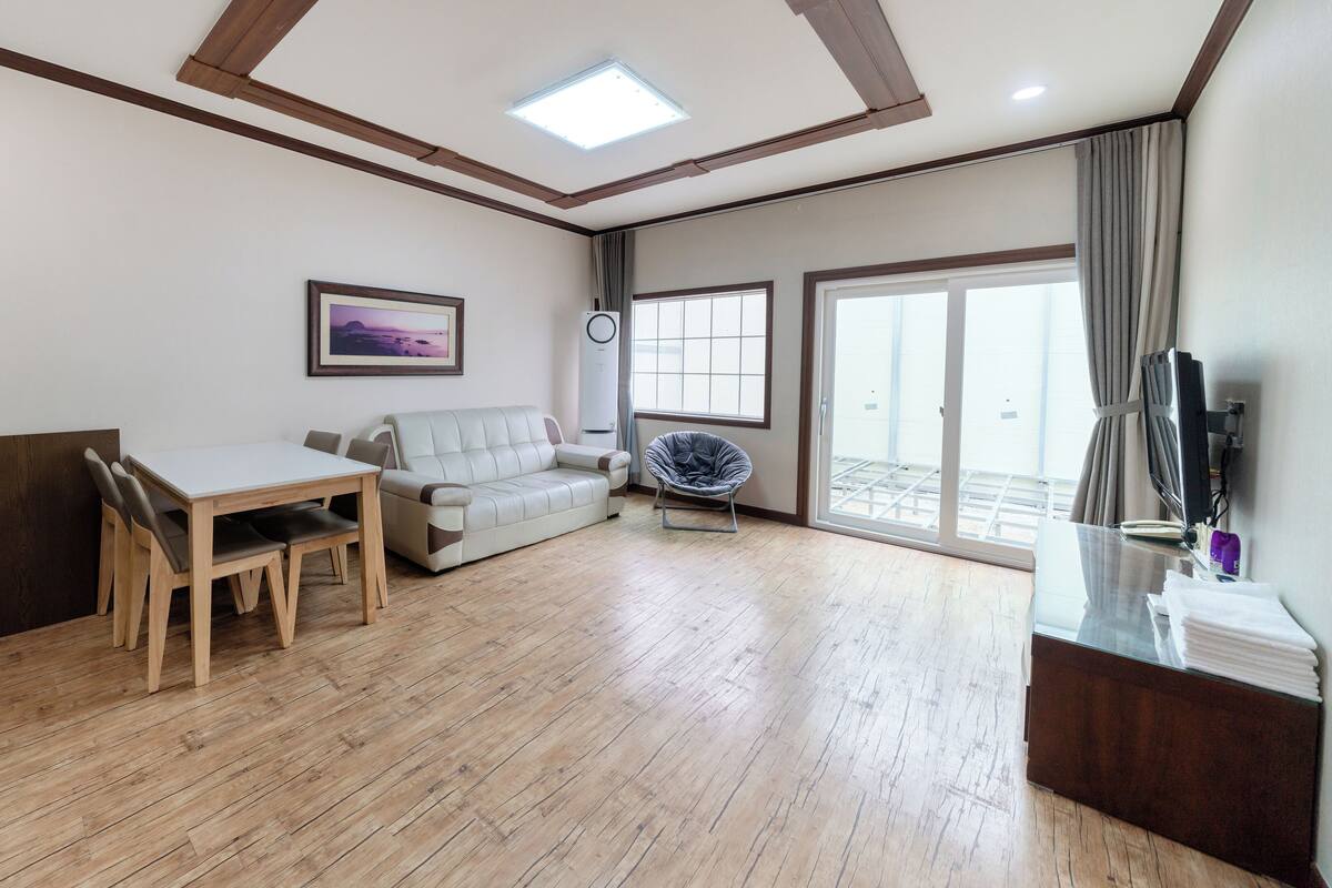 A spacious living area with a wooden floor, a sofa, a dining table with chairs, a ceiling fan, and a sliding glass door leading to a balcony.