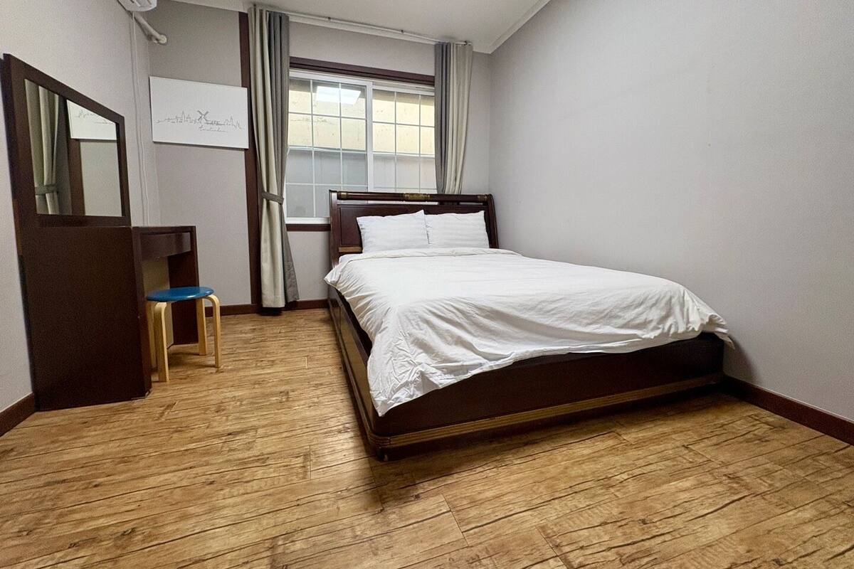 A bedroom with a wooden floor, a bed with white bedding, a desk with a chair, a window with curtains, and an air conditioning unit.