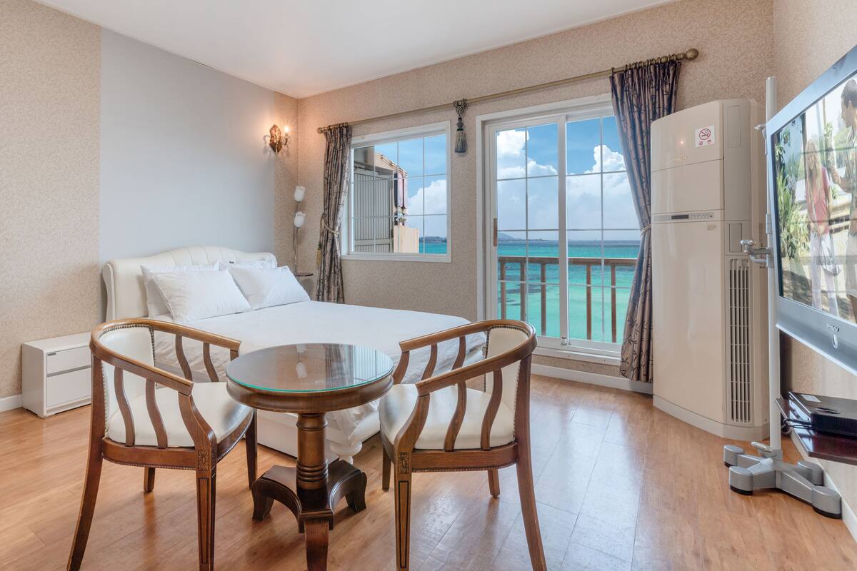 A hotel room with a bed, a desk, two chairs, a small table, a window with a view of the sea, and a balcony.