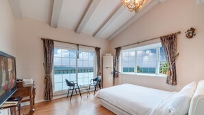 Un dormitorio con una cama grande, dos ventanas con vistas al océano, un escritorio con silla y una televisión.