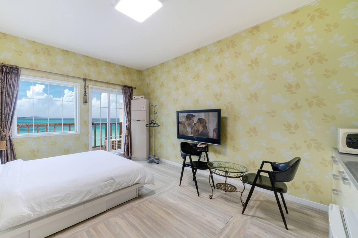 A hotel room with a bed, a TV, a desk, and a view of the sea.