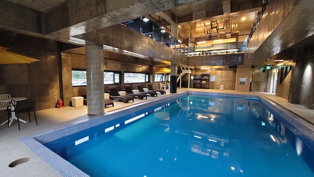 Una piscina interior con sillas de descanso, techo alto con vigas a la vista y vistas al exterior a través de grandes ventanales.