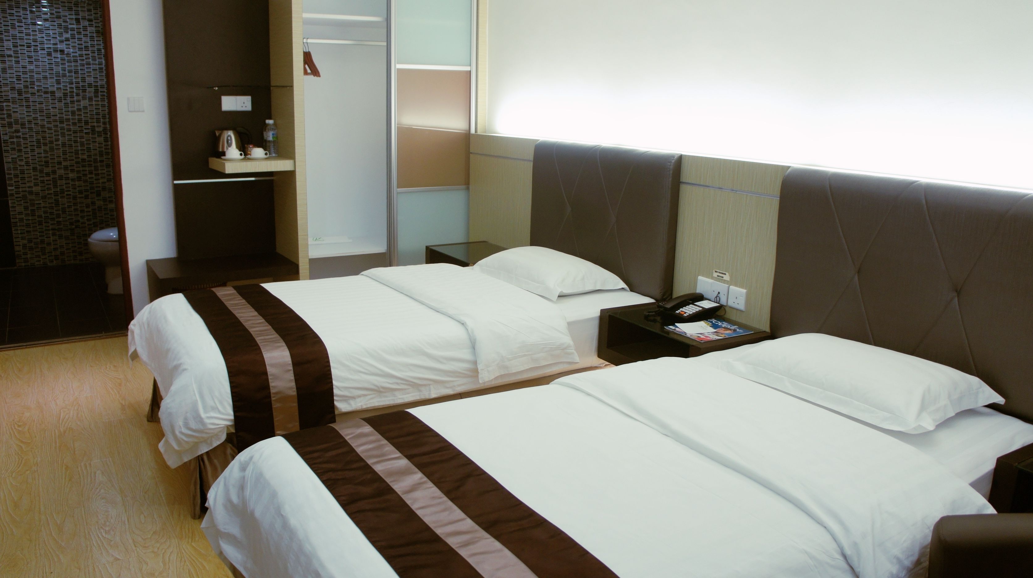 Une chambre d’hôtel avec deux lits, une table de chevet avec un téléphone et un magazine, une petite table avec une bouilloire et des tasses, et une salle de bain visible à travers une porte ouverte.