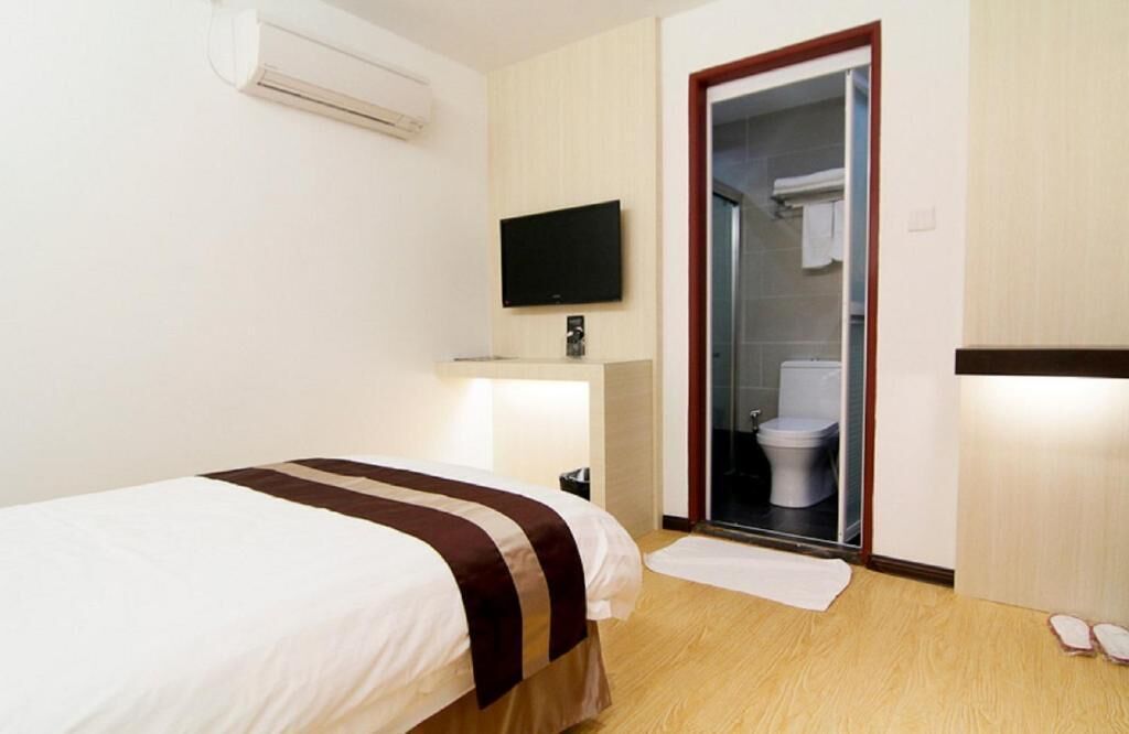 Quarto de hotel com cama, televisão, área de chuveiro e uma mesinha com abajur.