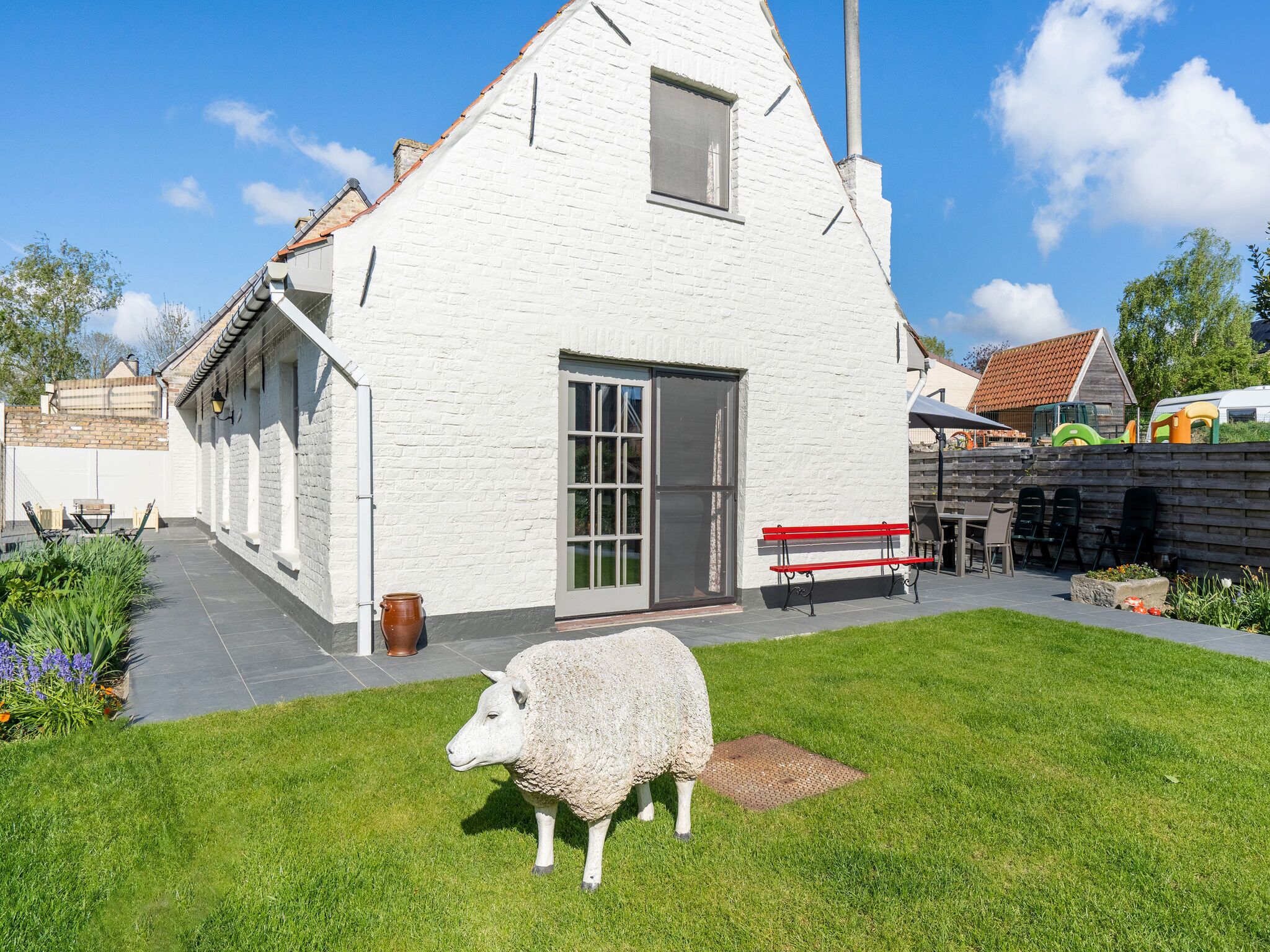 A white brick house with a large window and a door, a red bench, and a sheep statue in the yard.
