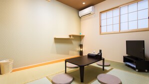 A traditional Japanese room with tatami flooring, a low table, cushions, a television, and an air conditioning unit.