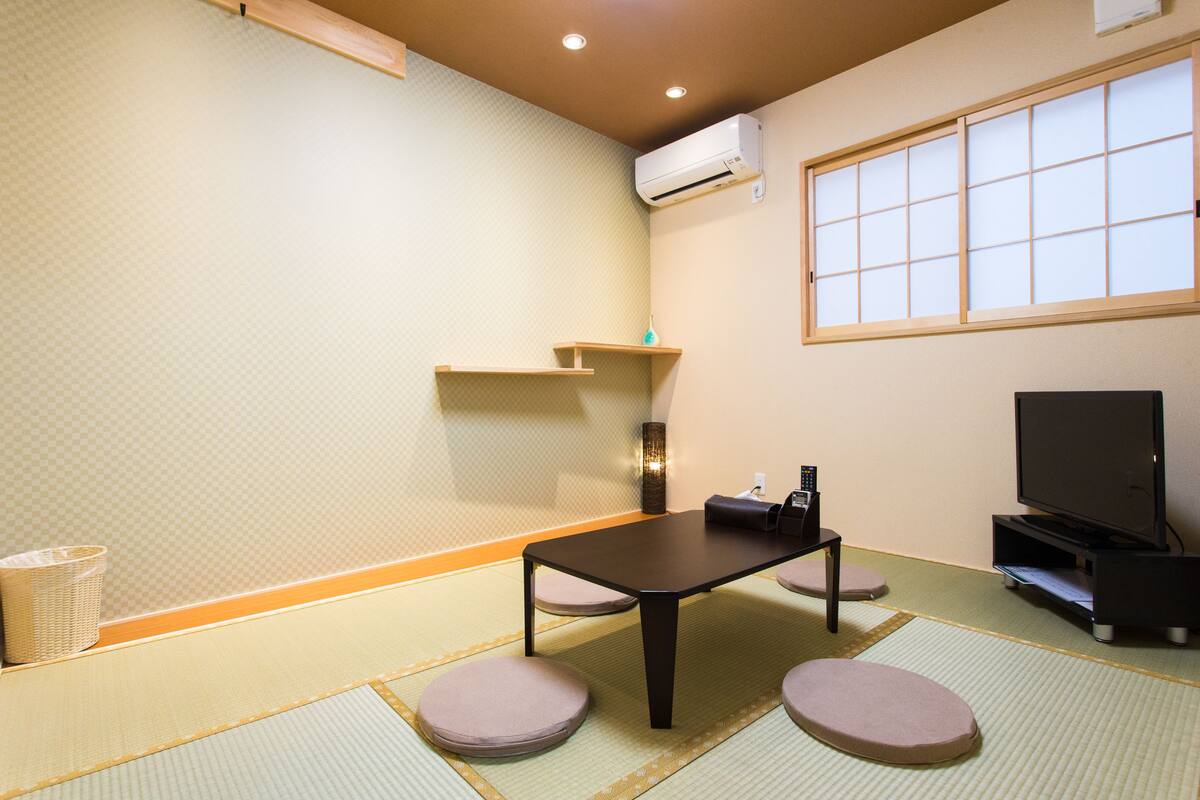 Una habitación japonesa tradicional con suelo de tatami, una mesa baja, cojines, televisor y equipo de aire acondicionado.