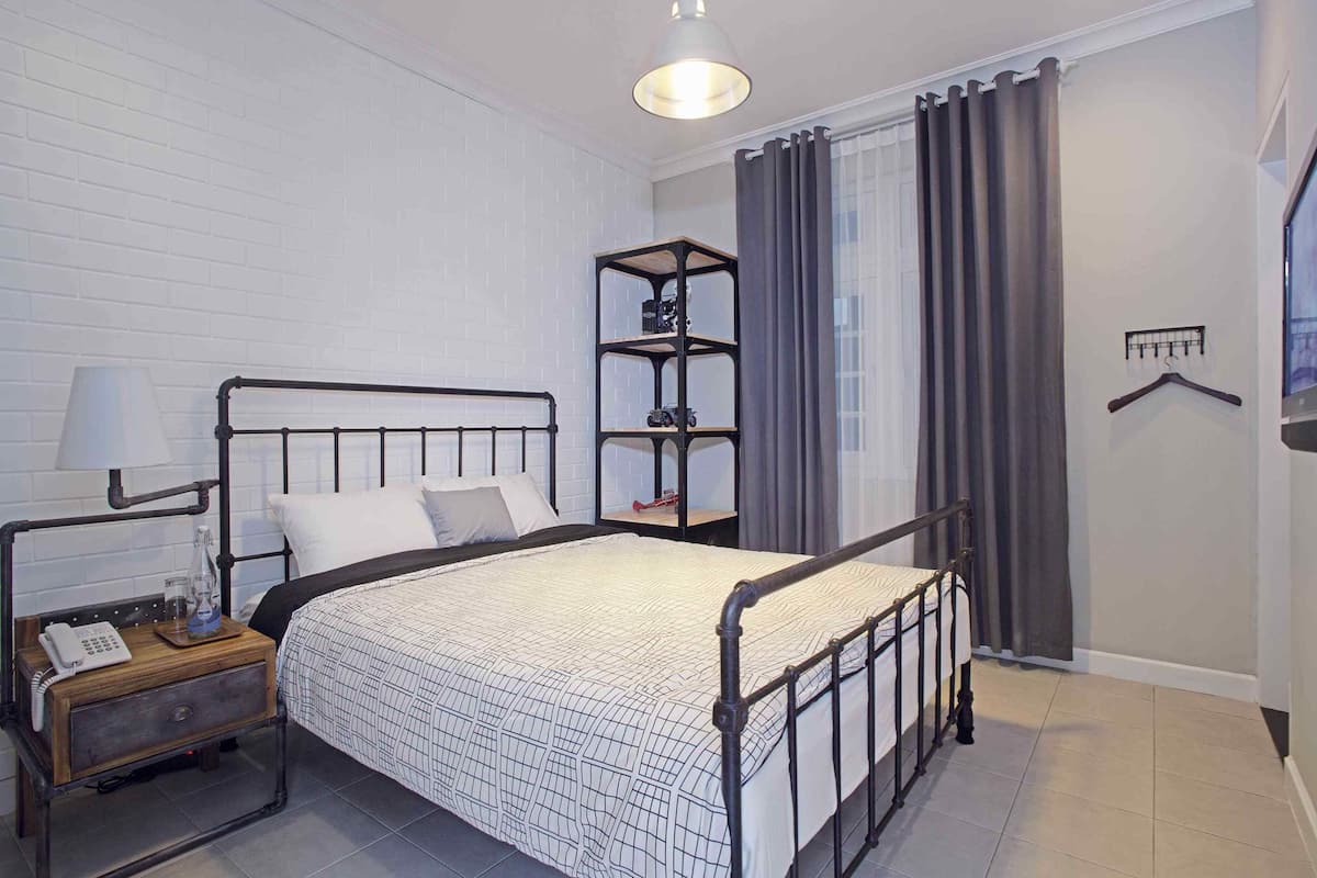 Une chambre à coucher bien rangée, avec un lit en métal, une table de chevet avec une lampe, une étagère et un portant à vêtements.