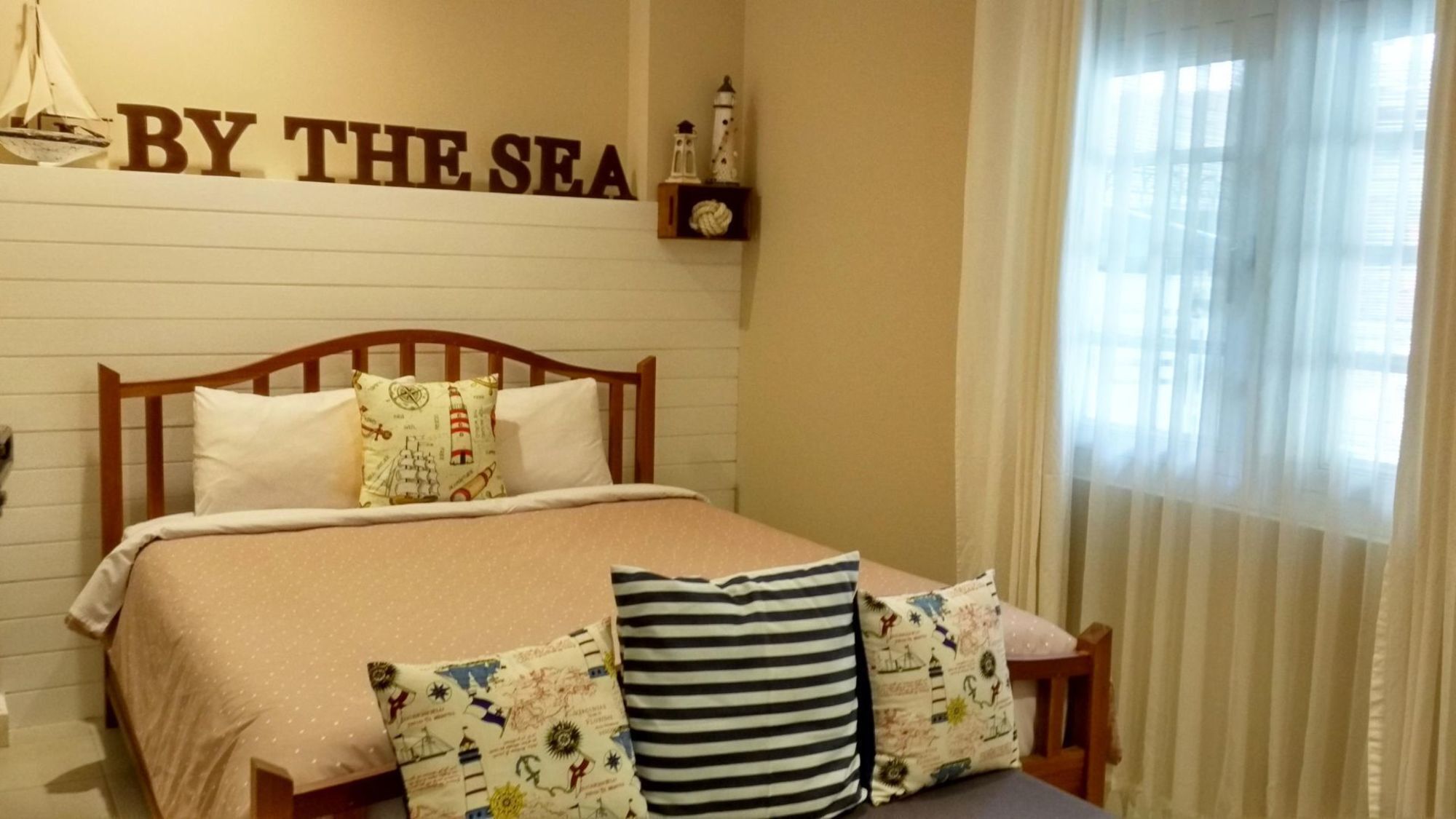 A bedroom with a bed, pillows, a headboard, a shelf with lighthouses, and a sign reading 'BY THE SEA'.