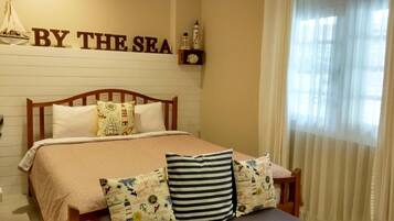 A bedroom with a bed, pillows, a shelf with decorative items, and a sign that reads "BY THE SEA".
