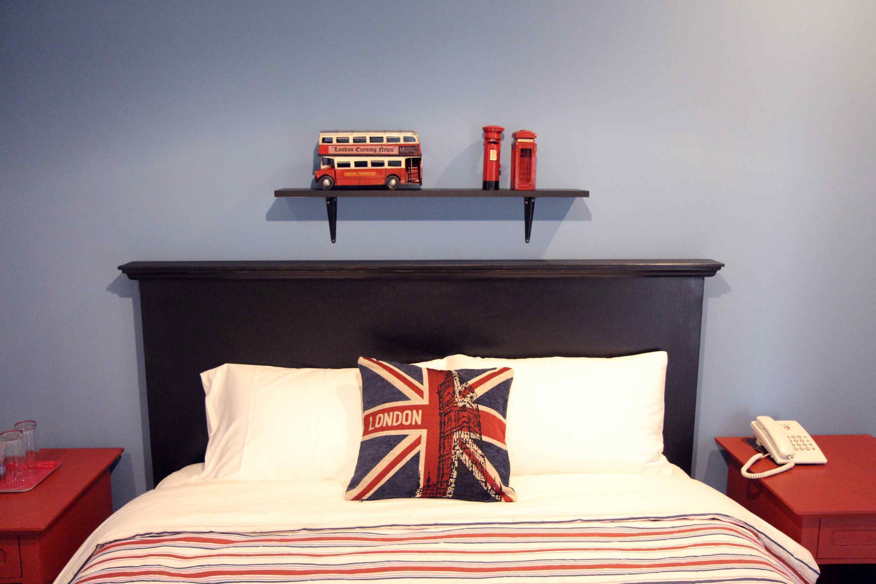 Una cama con una colcha a rayas, un almohadón con diseño de la bandera del Reino Unido, un estante con un autobús rojo de dos pisos y dos cabinas telefónicas rojas, y una mesita de noche con un teléfono blanco.