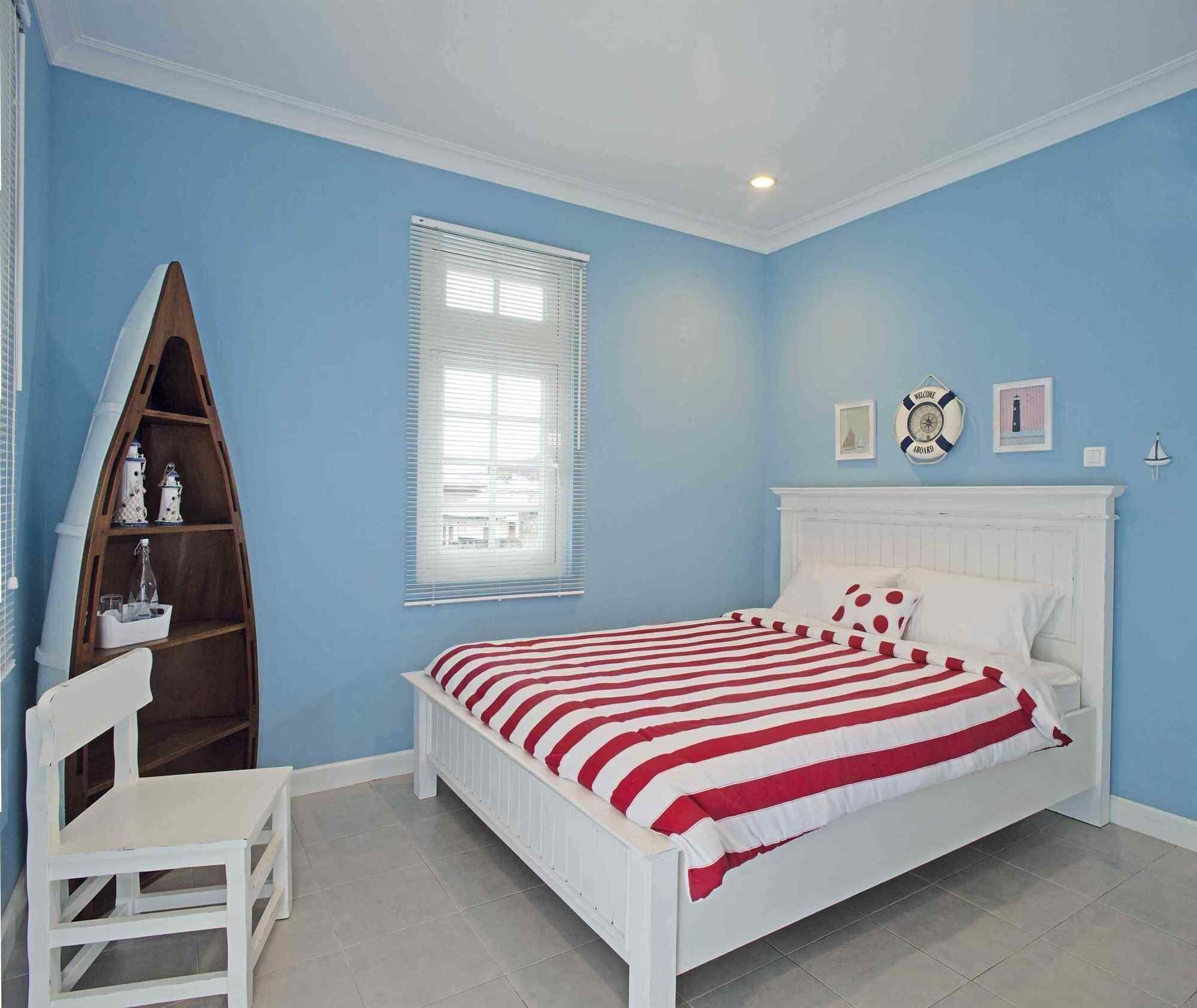 A bedroom with a bed, a white chair, a wooden shelf, a window with blinds, and a wall clock.