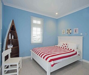 A bedroom with a bed, a white chair, a wooden shelf, a window with blinds, and a wall clock. - Cottonwood Bed & Breakfast House (Bandung)