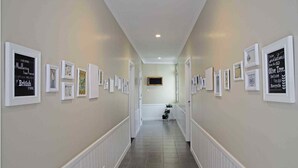 A hallway with framed pictures on the walls, a white door at the end, and a tiled floor. - Cottonwood Bed & Breakfast House (Bandung)