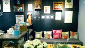 A cozy seating area with a dark wall adorned with various signs and decorations, including a sign that reads "Magic Happens Everyday" and another that says "Live Travel." There are pillows with text on them and a small shelf with a toy van. - Cottonwood Bed & Breakfast House (Bandung)