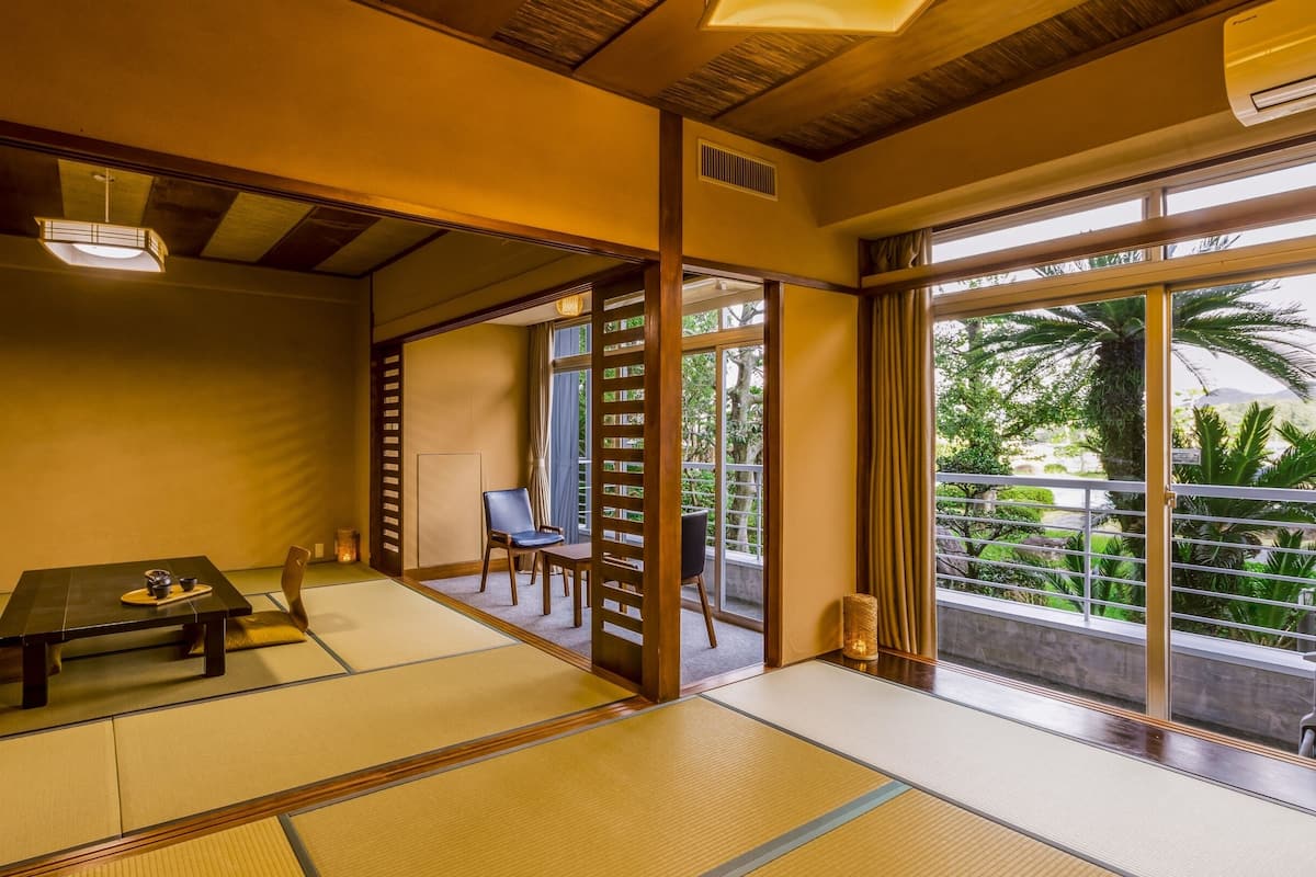A traditional Japanese room with tatami flooring, a low table with a tea set, sliding doors, and a view of greenery outside.