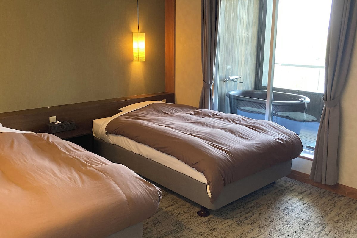 A hotel room with two beds, a nightstand, a lamp, and a view of the outside through a sliding door.
