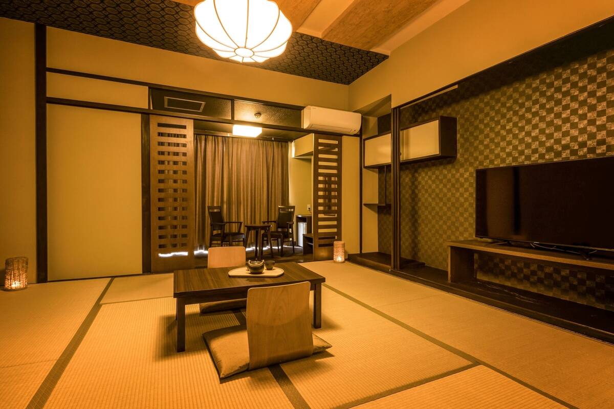 A traditional Japanese-style room with tatami flooring, a low table with cushions, a television on a stand, and a sliding door leading to another room.