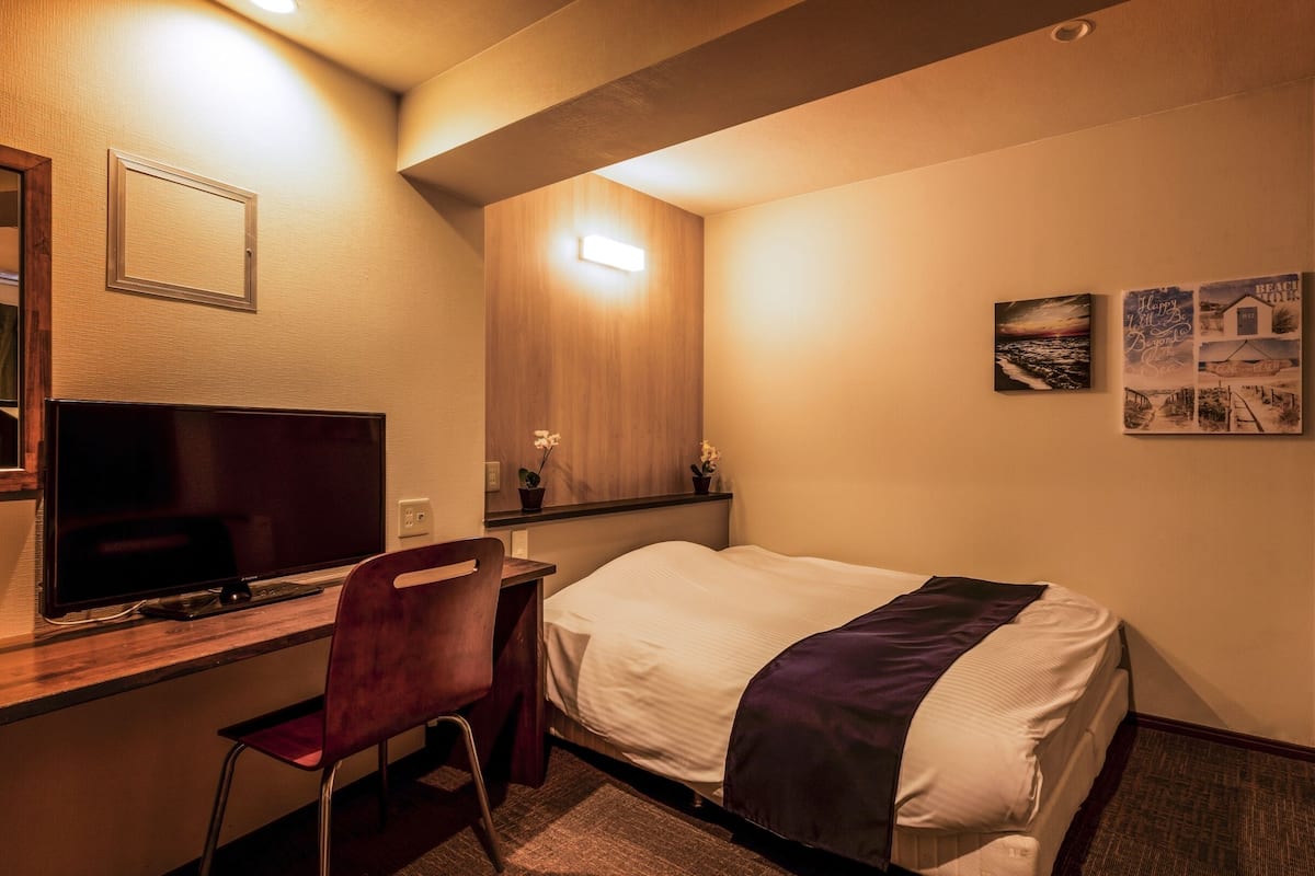 A hotel room with a bed, a desk with a TV, a chair, a small fridge, and wall art.