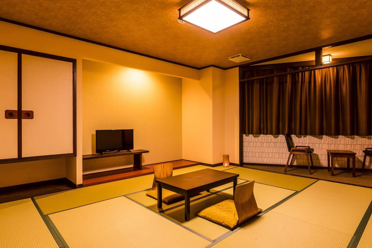 A traditional Japanese-style room with tatami flooring, a low table with a cushion, a television on a stand, and a sliding door.