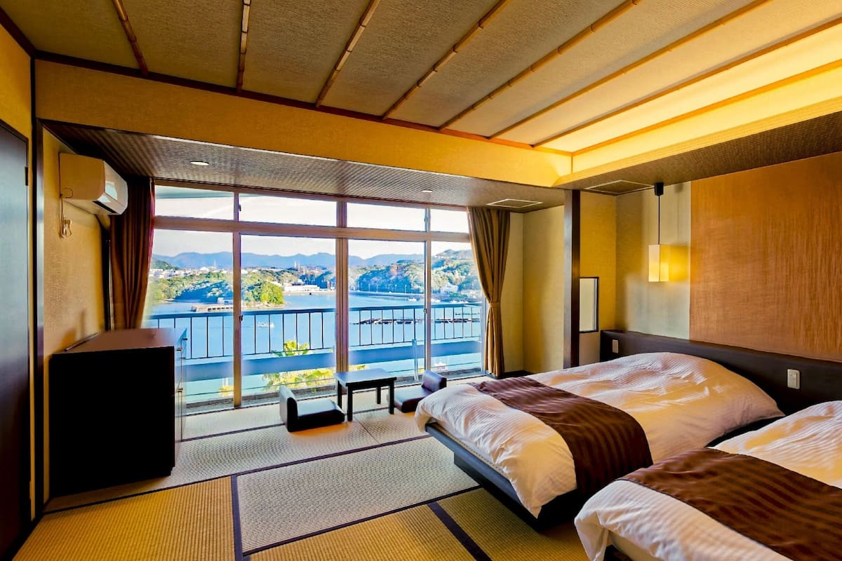 A hotel room with two beds, a large window with a view of a body of water and hills, and a small table with a chair.