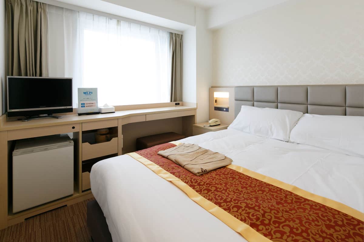 A hotel room with a bed, a desk with a TV, a mini-fridge, and a window with curtains.