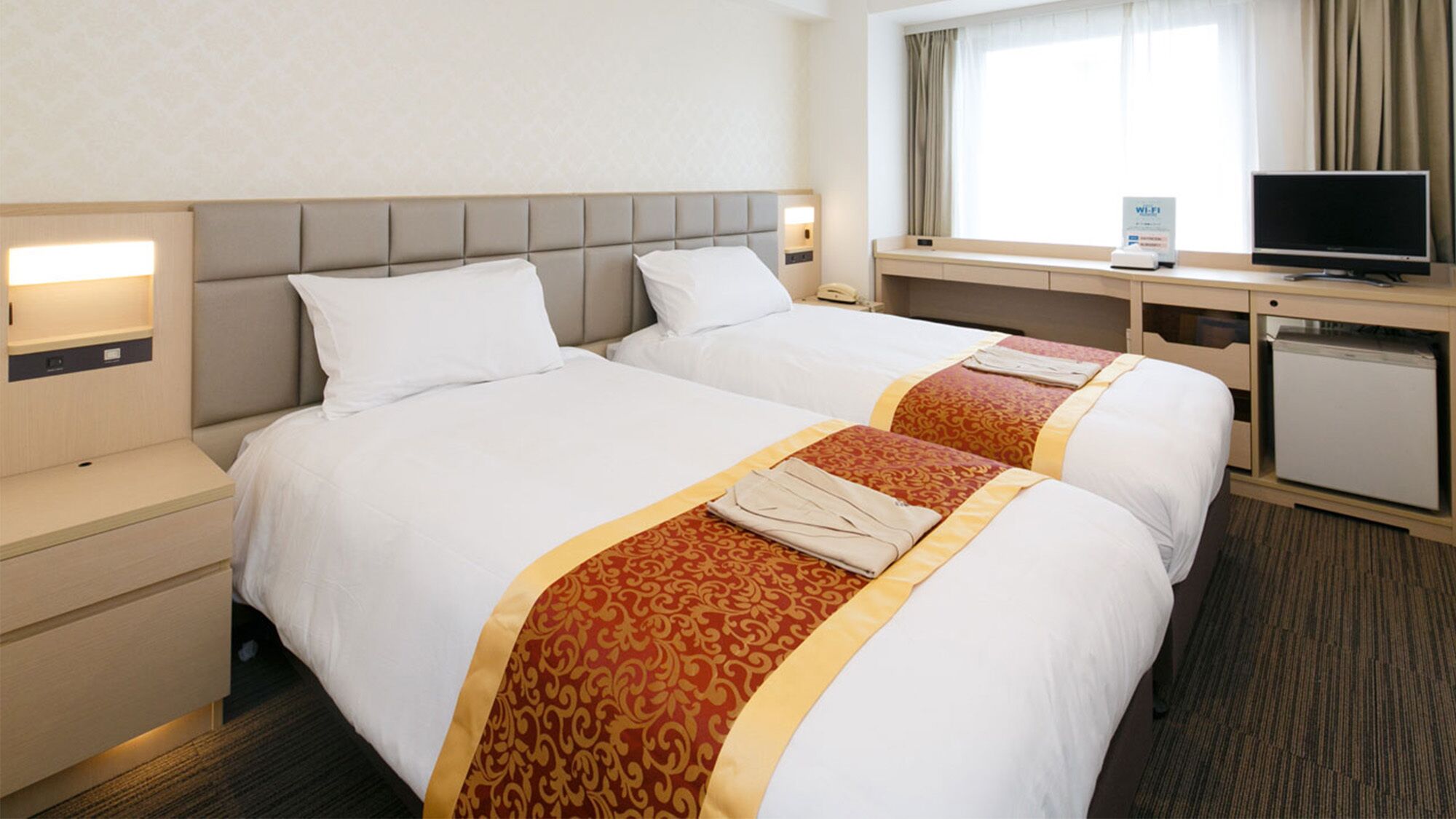 Ein Hotelzimmer mit zwei Betten, einem Schreibtisch, einem Fernseher und einem Kühlschrank.