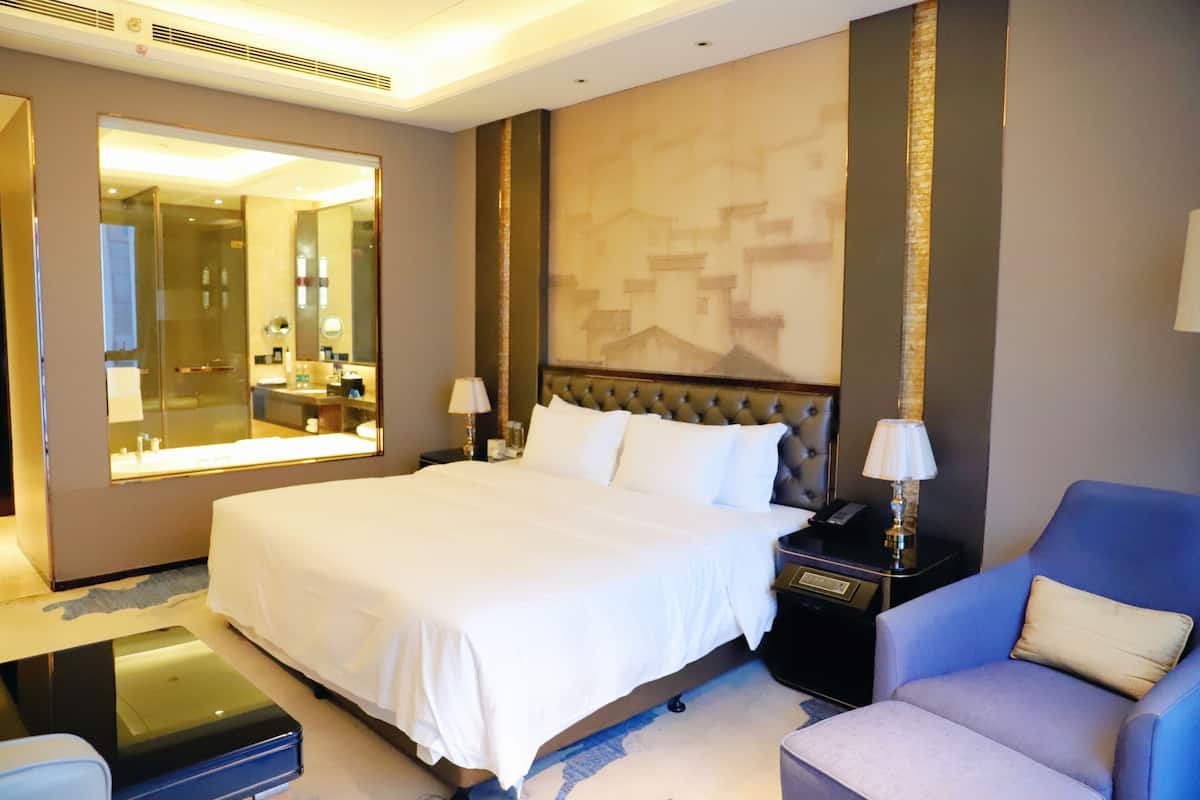 A hotel room with a large bed, a sitting area with a blue armchair, a side table with a lamp, and a large mirror reflecting the bathroom.