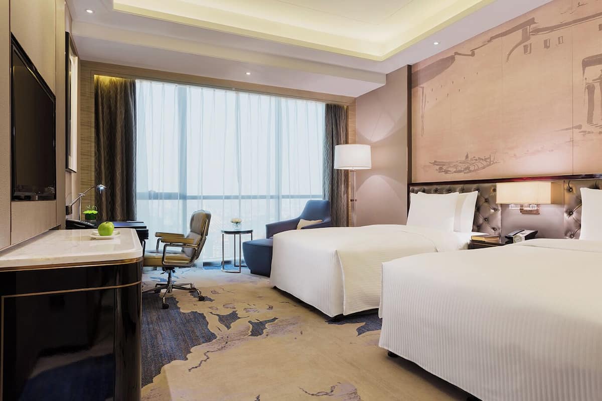 A hotel room with two beds, a desk with a chair, a TV, and a large window with curtains.