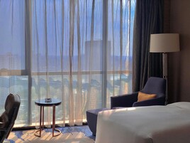 Habitación de hotel con un ventanal grande, una cama, una silla, una mesita y una lámpara.