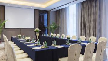 Un salón de conferencias con una larga mesa, sillas, una pantalla de proyector y un amplio ventanal con cortinas.