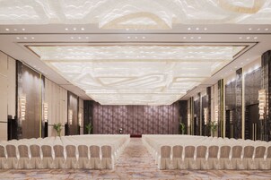 Un amplio vestíbulo de hotel con una larga recepción, una zona de estar amplia con filas de sillas y un techo decorativo con diseños de luz.
