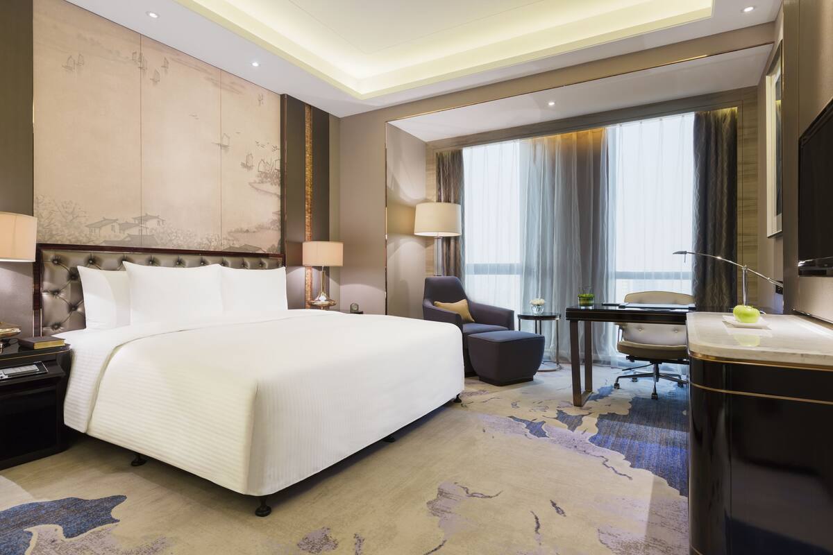A hotel room with a large bed, a desk, a chair, and a view of the city through the window.