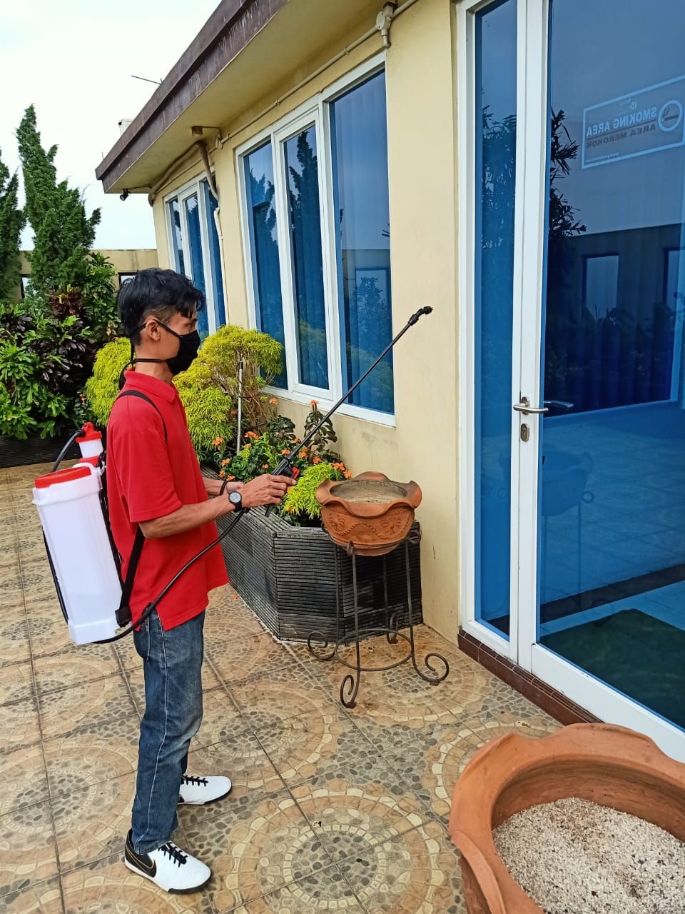 A person with a backpack spraying plants outside a building with blue-framed windows.